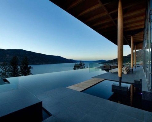 A modern home with a covered patio overlooks an infinity pool and a scenic lake surrounded by mountains at dusk. Glass railings and sleek furnishings add to the contemporary, tranquil atmosphere.