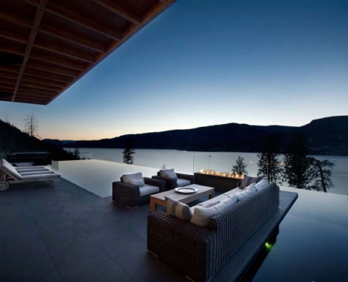 Modern outdoor patio with wicker sofas and a firepit beside an infinity pool, overlooking a tranquil lake and forested hills at dusk. The scene is calm with a clear sky as night approaches.