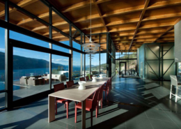 Modern dining area with red chairs and a wooden table, large floor-to-ceiling windows offering panoramic lake and mountain views, wood-beamed ceiling, pendant lights, and natural sunlight streaming inside.
