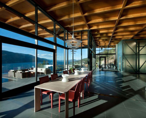 Modern dining area with red chairs and a wooden table, large floor-to-ceiling windows offering panoramic lake and mountain views, wood-beamed ceiling, pendant lights, and natural sunlight streaming inside.