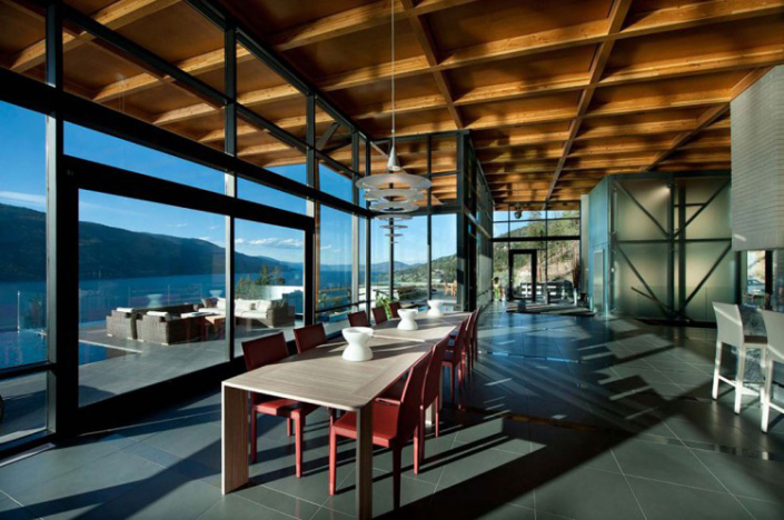 Modern dining area with red chairs and a wooden table, large floor-to-ceiling windows offering panoramic lake and mountain views, wood-beamed ceiling, pendant lights, and natural sunlight streaming inside.