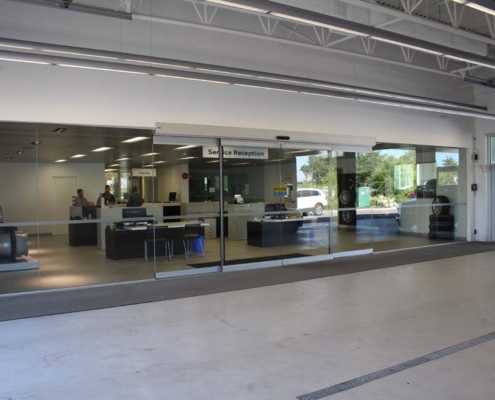 A car service center with a glass-walled service reception area. Inside, desks and chairs are visible, and a few people are at the counters. Outside, a white vehicle is parked near trees.
