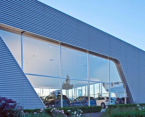 Modern car dealership building with large angled glass windows, showcasing several cars inside. The exterior features a geometric metal facade, landscaped bushes, and a clear blue sky in the background.