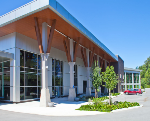 Modern building with large glass windows, wooden support beams, and a landscaped entrance. A small tree and green plants are near the entrance. A red car is parked in the lot under a clear blue sky.