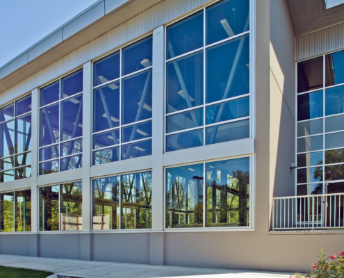 Modern building exterior with large, reflective glass windows and a metal railing. Trees and sky are reflected in the windows, and a concrete walkway runs alongside the building.