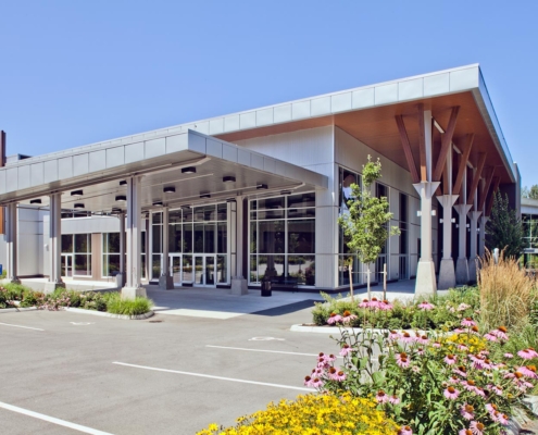 A modern building with a covered entrance, large glass windows, and wooden columns, surrounded by landscaped gardens with flowers and greenery under a clear blue sky.