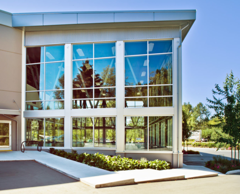 Modern building with large glass windows reflecting trees and sky. Entrance area has a concrete walkway, bike rack, small landscaped garden, and nearby parking spaces. Trees and greenery surround the building.