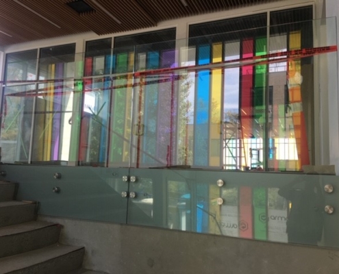 Glass panels with colorful vertical strips behind them line a modern interior stairway. Red caution tape is stretched across the top, and sunlight casts colored reflections onto the floor and walls.