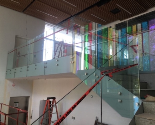A modern indoor staircase with glass railings and colorful vertical glass panels, marked off with red caution tape. A ladder and construction equipment are visible, indicating ongoing work.