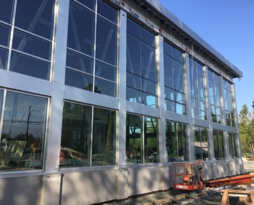 Close-up of a modern building under construction with large glass windows, metal framing, and construction equipment visible on the ground; trees and blue sky in the background.