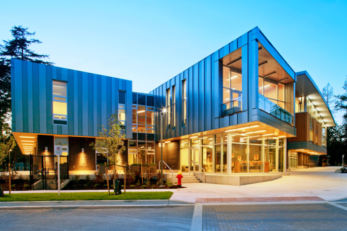 Modern building with large glass windows and blue metal panels, illuminated at dusk. Trees and landscaping surround the structure, and warm interior lights are visible through the windows. A red fire hydrant is at the curb.