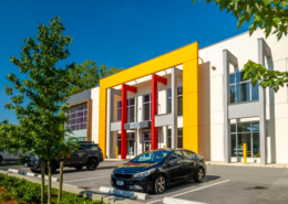 A modern office building with a bold yellow and red entrance facade, large windows, and parked cars in the lot on a sunny day. Trees and greenery are visible around the parking area.