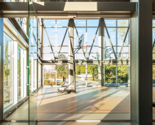 Indoor basketball court with glass walls and high ceilings, letting in abundant natural light. A basketball hoop is centered, and trees and greenery are visible outside through the large windows.