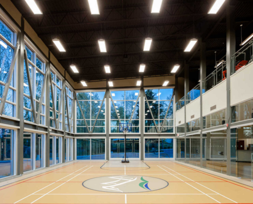 A modern indoor gymnasium with large windows, high ceilings, bright lighting, and a basketball hoop at the far end. The floor has sports court markings and a logo in the center. Trees and sky are visible outside.
