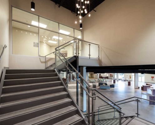 Modern interior staircase with metal railings leading to an upper floor, glass walls overlooking an open lobby area, and a contemporary chandelier with hanging globe lights above.
